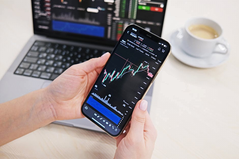 Close-up of hands holding smartphone displaying stock charts with laptop in the background on a desk.